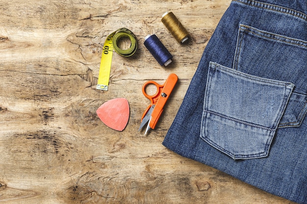 A person's hands carefully patching a hole in a pair of jeans with a colorful fabric scrap. Various sewing tools are laid out on a wooden table, highlighting the patching process.
