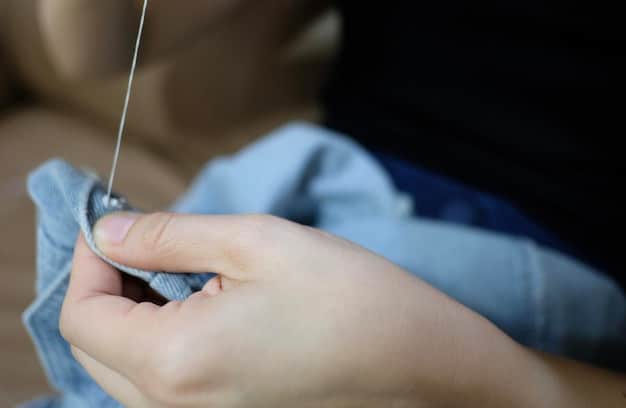 A hand holding a needle and thread, carefully sewing a button back onto a shirt. The focus is on the precision and care being taken during the repair process.