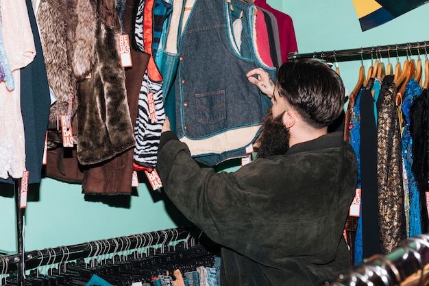 A person browsing through a well-organized rack of vintage clothing at a brightly lit thrift store in a trendy urban area. Other shoppers are visible in the background, also looking through the racks and trying on clothes.