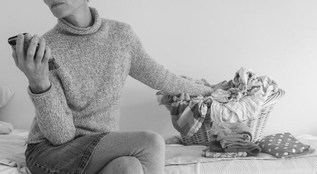 A woman showing a capsule wardrobe laid out on a bed, featuring versatile clothing items in neutral colors like black, white, and gray. Accessories like scarves and jewelry are also included.