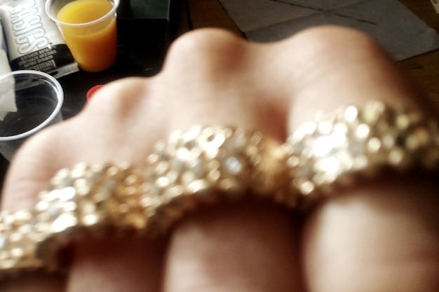 A close-up shot of a woman's hand adorned with multiple delicate gold rings, showcasing various textures and designs, set against a blurred background of a stylishly decorated room.