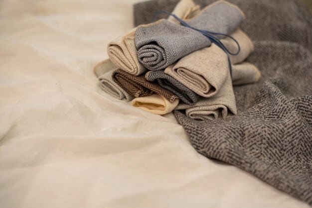 A close-up shot of a neatly arranged pile of clothing in neutral colors, including a white blouse, dark jeans, a gray sweater, and a black blazer. The clothing is folded or rolled tightly to save space, and a packing cube is visible in the background.