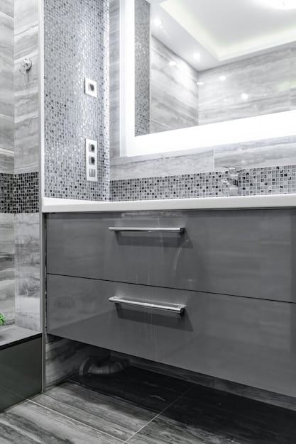 Close-up of a bathroom vanity with a faux-marble countertop, ceramic tile backsplash, and stylish brushed nickel faucet. There's a small, decorative soap dispenser and folded hand towels on the counter. The lighting is warm, highlighting the details of the materials.