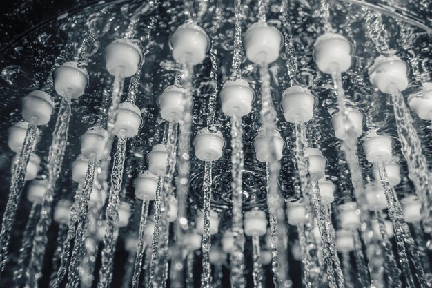 A close-up of a modern showerhead with a rainfall effect, installed in a tiled shower stall. The tiles are a light gray color, and the showerhead is chrome. Water droplets are visible, showcasing the showerhead in action.
